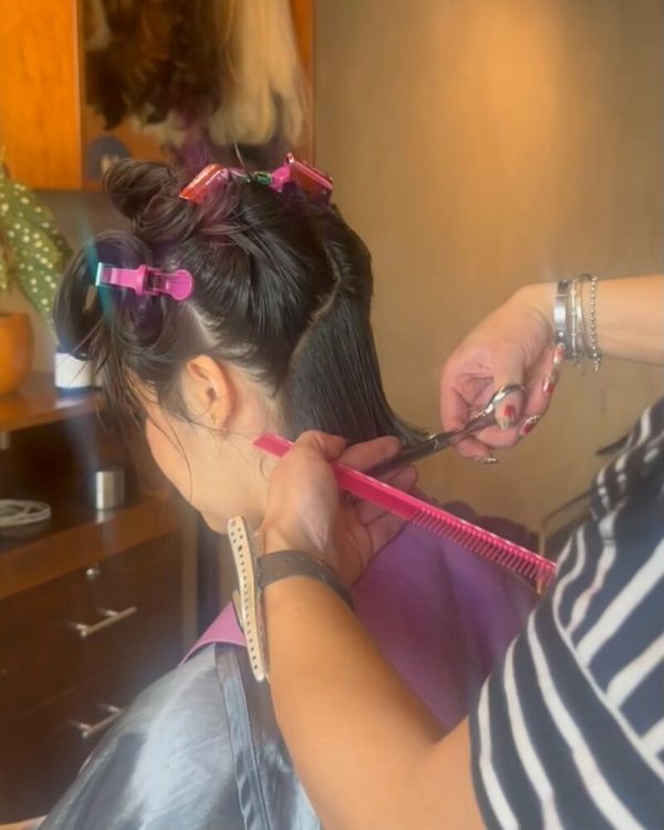 A woman sits in a salon chair as a hairdresser styles her hair with a brush and blow dryer.