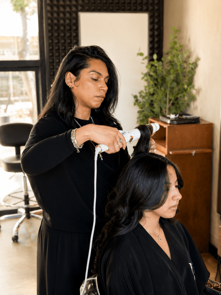 A hairstylist uses a curling iron on a client's hair in a well-lit salon. The scene highlights professional hair care and styling techniques.
